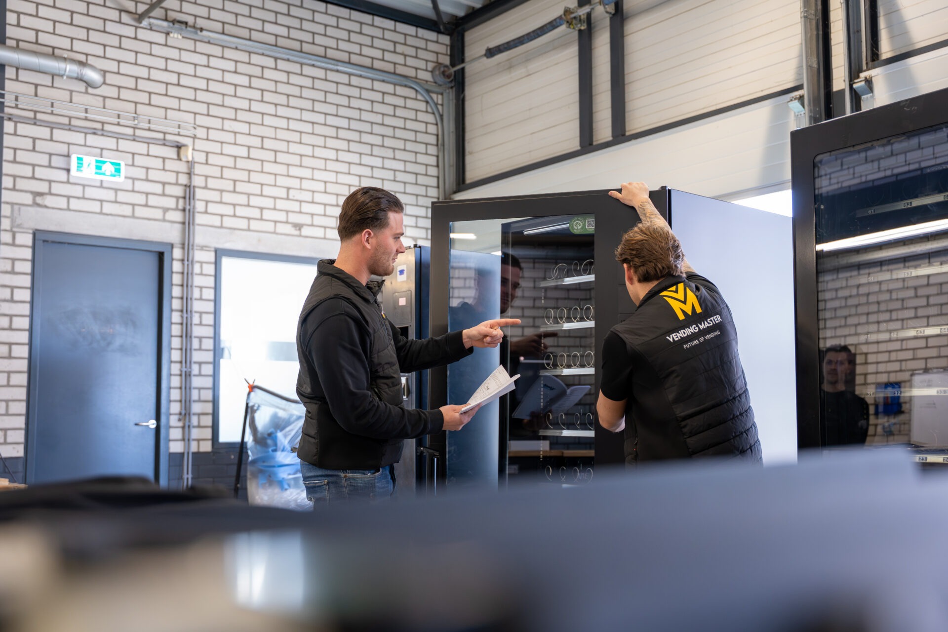Twee monteurs bij een vendingmachine in de werkplaats waarbij een monteur uitleg en aanwijzingen geeft aan een andere monteur over onderhoud en techniek van de automaat