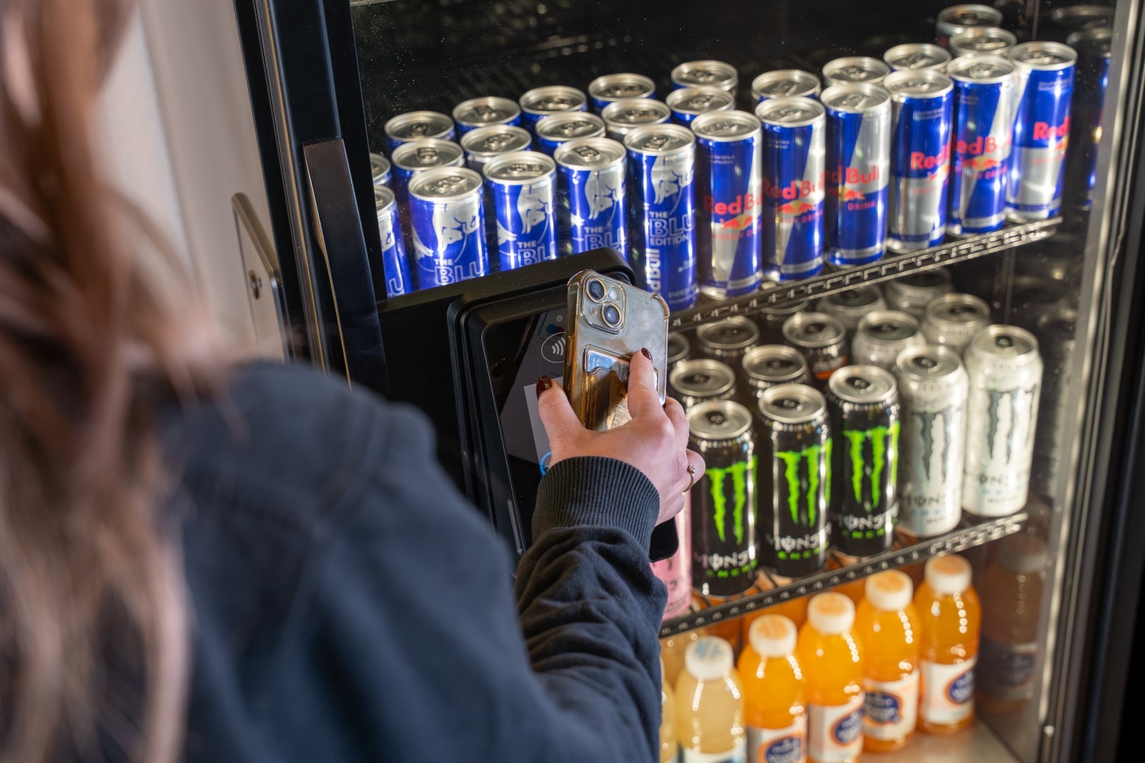 Een persoon die contactloos betaalt met een smartphone bij een Vending Master frisdrankautomaat gevuld met blikjes Red Bull en Monster Energy.
