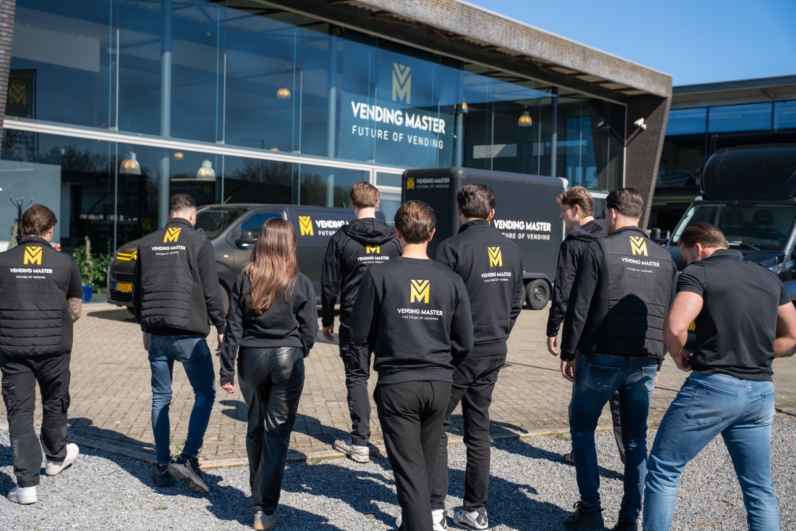 Een groep medewerkers van Vending Master, van de achterkant gefotografeerd in zwarte bedrijfskleding met logo, lopend richting het hoofdkantoor waar ook een bedrijfswagen en aanhanger met branding geparkeerd staan.