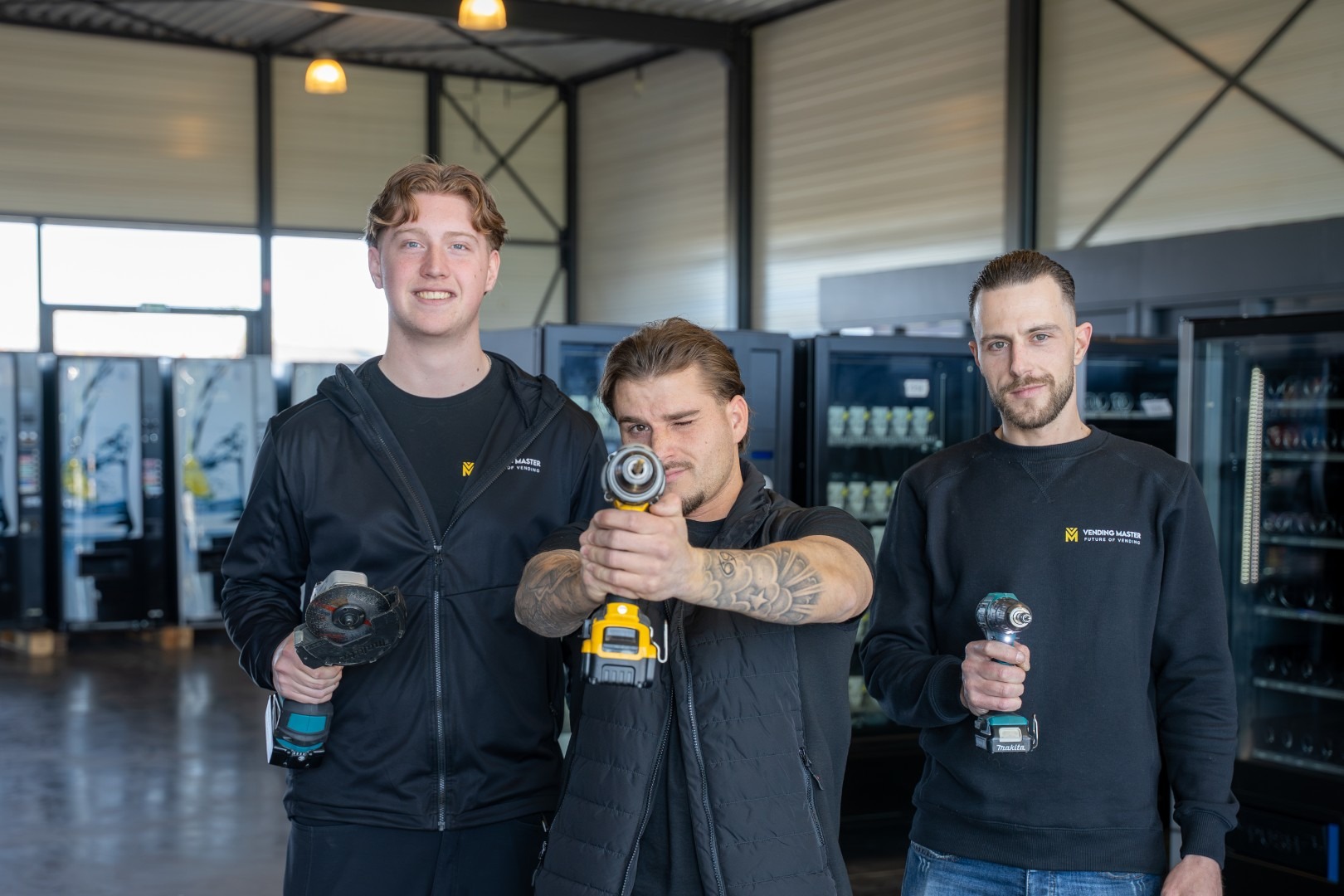 Drie medewerkers van Vending Master in het magazijn die poserend gereedschap vasthouden, zoals een boormachine en slijptol, met op de achtergrond diverse vending machines.